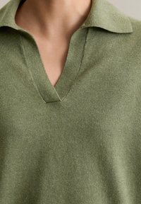 Sweter w serek z miękkiej, teksturowanej, oliwkowozielonej tkaniny. Posiada klasyczny kołnierz i subtelne przeszycia, które podkreślają proste, uniwersalne detale wzornicze.