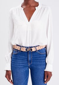 Blouse blanche avec un col à volants et des détails subtils en fil brillant, associée à un jean bleu taille haute et une ceinture élastique beige avec des accessoires dorés.