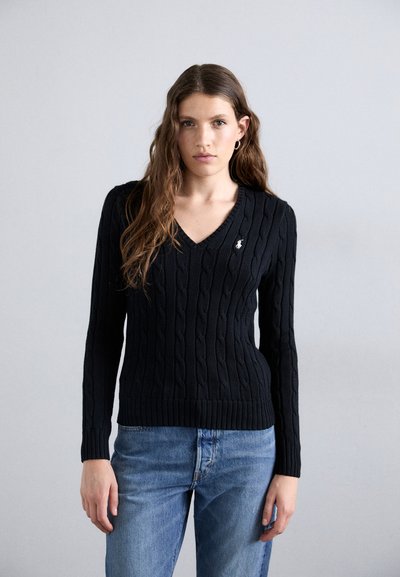 Suéter negro de punto trenzado con escote en V, puños y dobladillo acanalados. Presenta un pequeño emblema en el lado izquierdo del pecho. Combinado con jeans azules.