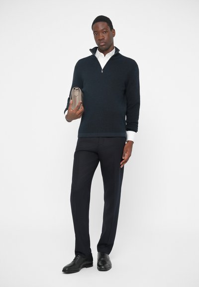 Pullover de tricot bleumarin cu guler cu fermoar pe jumătate, purtat peste o cămașă albă. Pantaloni închiși la culoare și pantofi negri. Ține un pouch texturat gri.