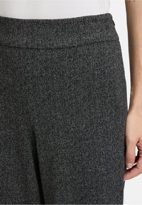 Gros plan sur un pantalon gris texturé avec un motif de chevrons subtil, porté par une personne avec un haut blanc, montrant la taille et la main droite.