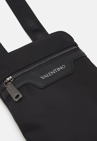 Sacoche noire en nylon avec une poche zippée, tissu texturé et logo "Valentino" en relief en haut. Sangles ajustables incluses.