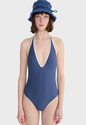Młoda kobieta ubrana w niebieski prążkowany jednoczęściowy kostium kąpielowy z białą lamówką i niebieskim kapeluszem typu bucket, stojąca na tle jasnego, jednolitego tła.