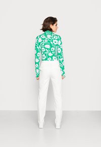 Turtleneck de manga longa em verde com grandes padrões florais brancos, combinado com calças brancas. Textura suave, design ajustado.