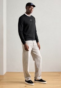 Felpa nera con logo, jeans grigi chiari e sneaker nere con suola bianca; presenta un cappellino nero con accento blu sul logo.