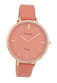 Montre en or rose avec un cadran rond pêche, des marqueurs d'heure minimalistes et un bracelet en cuir souple pêche assorti. "Vintage" imprimé sur le cadran.