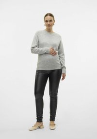 Lichtgrijze gebreide trui met lange mouwen, gecombineerd met zwarte neplederen leggings en beige hakken, staand tegen een eenvoudige achtergrond.