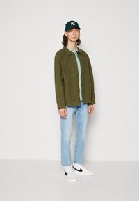 Grønn jakke med knappelukking over en lyseblå skjorte, kombinert med lyseblå jeans og hvite joggesko med en svart swoosh-logo.