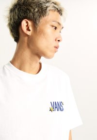 White cotton t-shirt featuring blue "VANS" logo with gold star accents, round neckline, and short sleeves. Soft texture, casual fit.