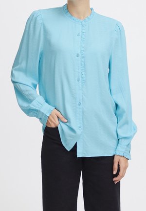 Lyseblå skjorte med knaplukning og rund hals, med rynket front og lange ærmer. Stoffet har en let struktureret overflade. Bæres over sorte bukser.
