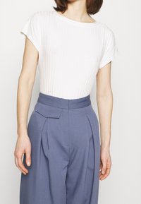 Donna che indossa un top bianco a righe con maniche corte, infilato in pantaloni blu a vita alta con pieghe e una tasca a patta sul lato destro.
