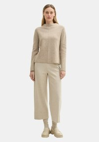 Beige Strickpullover mit hohem Kragen, kombiniert mit weiten beigefarbenen Hosen. Das Model trägt hellfarbene, robuste Slipper.