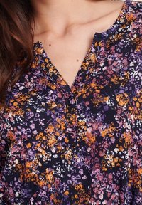 Halenka s květinovým vzorem v tmavě námořnické barvě se oranžovými, fialovými a bílými akcenty. Texturovaná látka má mírně uvolněný střih a výstřih do V.