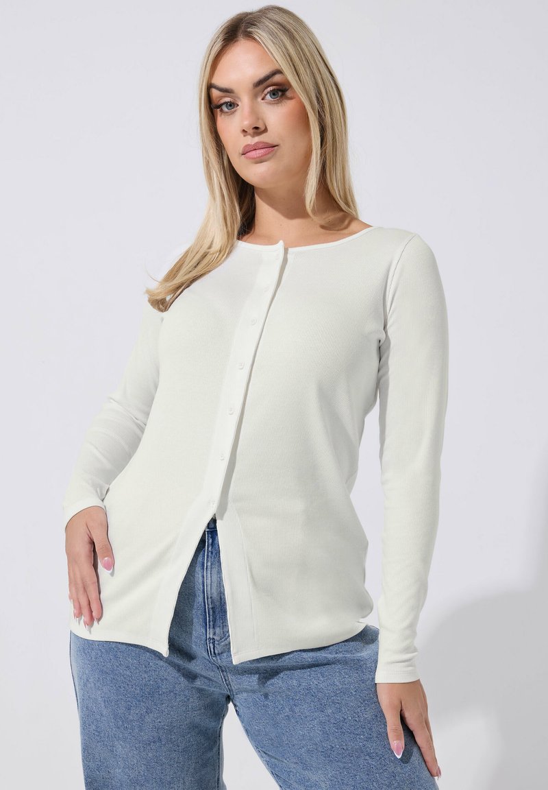 Blonde Frau, die eine weiße, langärmlige, geknöpfte Strickjacke und blaue Jeans trägt, mit einer Hand in der Hüfte vor einem einfarbigen Hintergrund posiert.