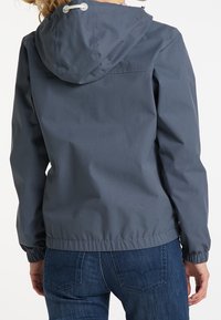 Veste imperméable grise avec capuche, poignets élastiques et taille cintrée ; texture lisse et design minimaliste sans motifs visibles.