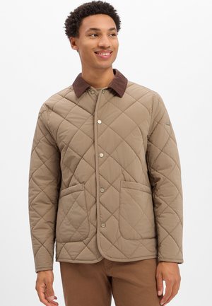 Mann trägt eine hellbraune gesteppte Jacke mit großen Vordertaschen und einem dunkelbraunen Kragen, lächelt und schaut nach rechts.