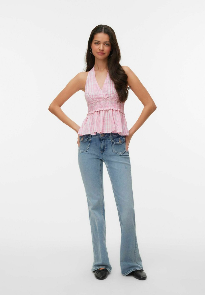 Femme aux longs cheveux foncés portant un haut dos nu rose et un jean évasé bleu clair, debout les mains sur les hanches sur fond blanc uni.