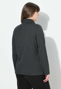 Donna con capelli castani lisci che indossa una giacca lunga a maniche lunghe scura con un motivo e pantaloni neri texturizzati, rivolta di lato su uno sfondo bianco.
