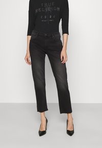Jeans de mezclilla negra con corte recto y dobladillo sin terminar. Combinados con una camiseta negra de manga larga con gráficos de texto brillantes. Tacones altos negros.