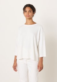 Ung kvinne med krøllete hår iført en løs hvit langermet topp og hvite bukser, stående mot en ensfarget lys bakgrunn.