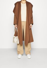 Brun ullblandad omslagsoverall över en krämfärgad tröja, i kombination med beige byxor och vita sneakers. Tillbehör inkluderar en vävd väska.