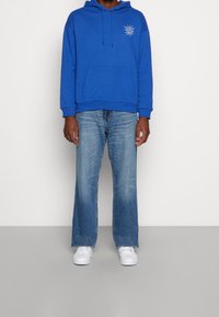 Sweat à capuche bleu avec une poche avant et un motif de soleil, associé à un jean évasé bleu clair et des baskets blanches. Tissu doux, coupe décontractée.