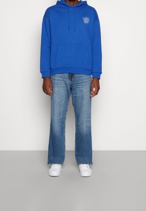 Blauwe hoodie met een voorvak en een zonafbeelding, gecombineerd met lichtblauwe uitlopende jeans en witte sneakers. Zachte stof, casual pasvorm.
