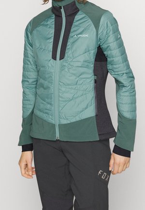 Persoon draagt een groene en grijze Vaude ritsjack en zwarte Fox-broek, staat met de armen ontspannen langs het lichaam.