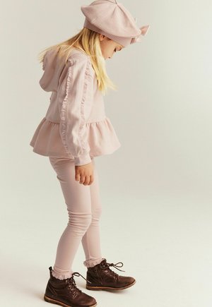 Jong meisje in roze outfit en baret kijkt omlaag, staat met handen langs haar zij en draagt bruine veterlaarzen.