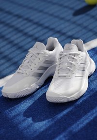 Pair of white athletic shoes with laces on a blue tennis court near a white boundary line and a yellow tennis ball in background.