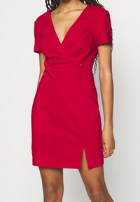 Robe rouge avec manches courtes, décolleté en V, design ajusté, dotée de fentes latérales. Fabriquée en tissu lisse avec des accents matériels minimalistes.