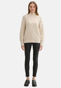 Světle béžový svetr s vysokým límcem vyrobený z texturovaného materiálu, kombinovaný s černými skinny kalhotami a robustními černými botami.