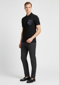 Černé polo triko s texturovanou džínovou kapsou, kombinované s tmavými úzkými kalhotami a černými mokasíny, na neutrálním pozadí.