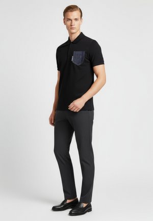 Czarna koszulka polo z fakturowaną kieszenią z denimu, w zestawieniu z ciemnymi, dopasowanymi spodniami i czarnymi mokasynami, na neutralnym tle.
