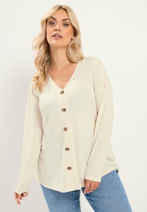 Blonde Frau trägt ein cremefarbenes, langärmliges, geknöpftes Oberteil und blaue Jeans, steht vor einem einfarbigen hellen Hintergrund.
