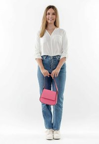 Bolso cruzado sintético rosa con tapa, contrarrestado por bordes negros, cuenta con una correa corta. El modelo lleva una camiseta blanca y jeans azules.