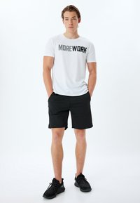 Camiseta blanca de manga corta con el texto negro "MOREWORK", combinada con pantalones cortos negros y zapatillas deportivas negras.