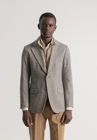 Blazer de cuadros grises con un diseño estructurado, solapas con muesca, cierre de un solo botón y dos bolsillos frontales, llevado sobre una camisa beige.