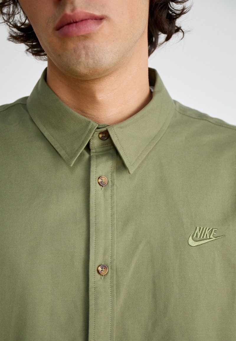 Camisa de algodón verde oliva con diseño de botones, que presenta un cuello con puntas, botones de metal y un logo de Nike bordado en el lado izquierdo del pecho.