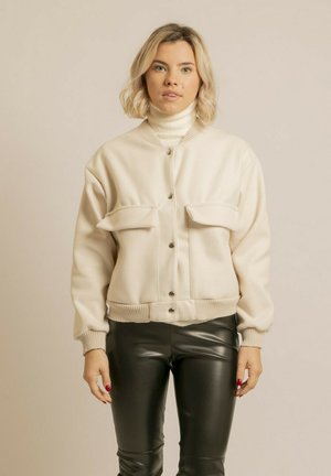 Jeune femme portant une veste crème boutonnée sur un col roulé blanc et un pantalon en cuir noir, debout devant un fond beige uni.