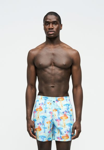 Γυμνασμένος άνδρας στέκεται μπροστά σε απλό φόντο, φορώντας πολύχρωμα μαγιό με μοτίβα πορτοκαλί, μπλε και ροζ μέδουσες και με κορδόνι στη μέση.
