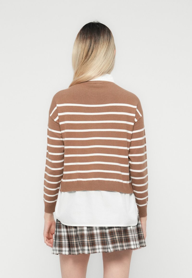 Maglione a righe marroni e bianche con design svasato, indossato sopra una camicia bianca, abbinato a una gonna a quadri con tonalità di marrone e nero.