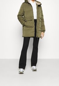 Person wearing an olive green padded coat, white sweater, black flared pants, and white sneakers, standing against a plain white background.