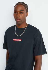 Un jeune homme avec des cheveux tressés portant un T-shirt Quiksilver noir et un collier en chaîne argenté sur un fond blanc uni.