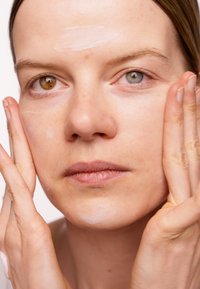 Hautpflegeanwendung mit einer leichten Creme auf einem blassen Gesicht. Merkmale ungleicher Augenfarben: ein braunes, ein blau-grünes Auge. Sichtbare Hautstruktur.