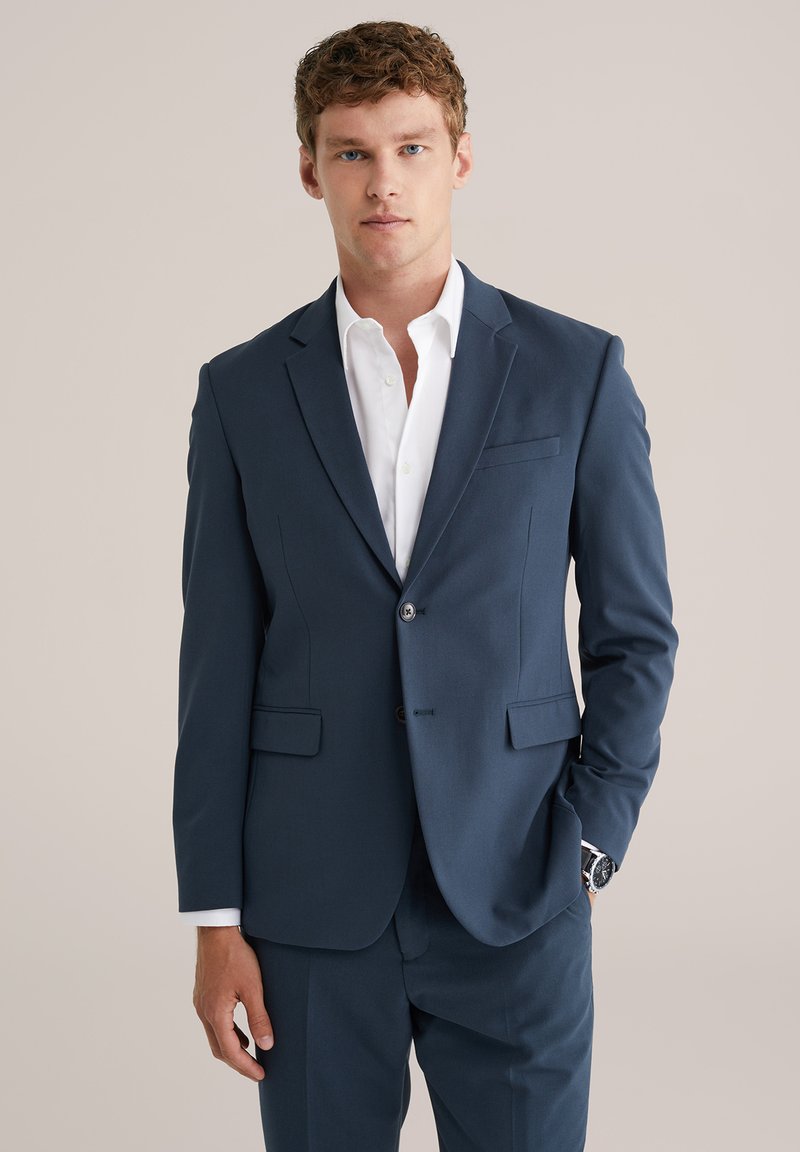 Navyn blå kostymjacka med notch-lapel, två framfickor och enkelknäppning, i kombination med en vit skjorta. Slät textur.