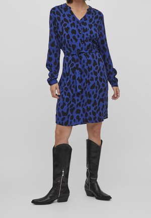Robe bleu imprimé léopard avec un décolleté en V, des manches longues et une ceinture à la taille ; associée à des bottes de cowboy noires jusqu'aux genoux avec des zips.