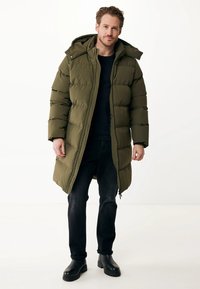 Mand iført en olivengrøn lang pufferjakke med hætte, sorte jeans, sorte støvler og en mørk skjorte, stående mod en hvid baggrund.