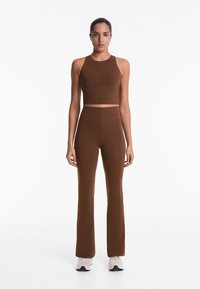 Braunes Sportoberteil mit hohem Kragen, kombiniert mit passenden ausladenden Leggings aus glattem Material. Getragen mit hellen Turnschuhen.