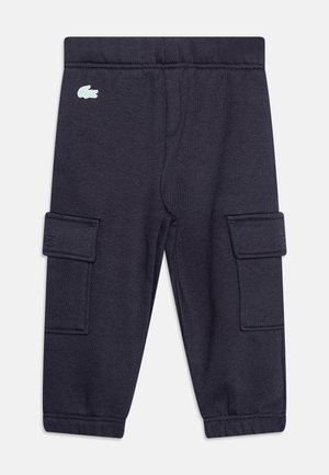 Enfant portant une veste zippée Lacoste gris clair, un pantalon cargo jogging foncé et des baskets blanches avec des lacets noirs, debout les mains dans les poches.
